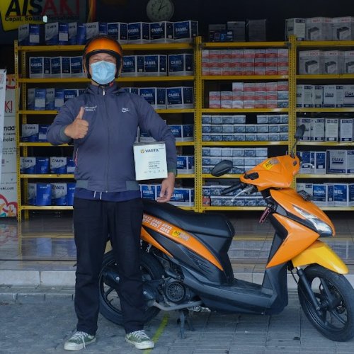 Seseorang sedang memegang aki dan di belakangnya ada toko aki.