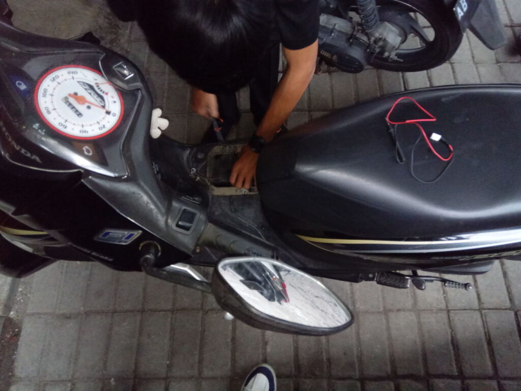Teknisi sedang memasang aki pada motor matic dengan jok terbuka di lokasi outdoor, menggunakan peralatan sederhana.