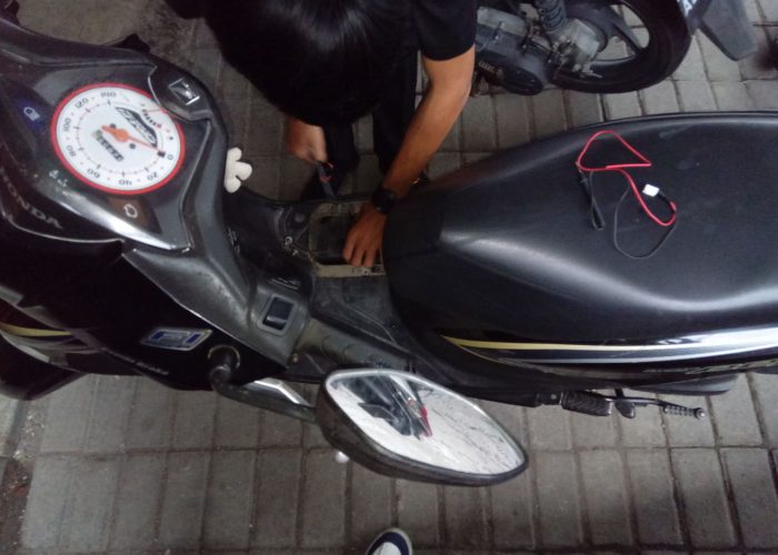 Teknisi sedang memasang aki pada motor matic dengan jok terbuka di lokasi outdoor, menggunakan peralatan sederhana.